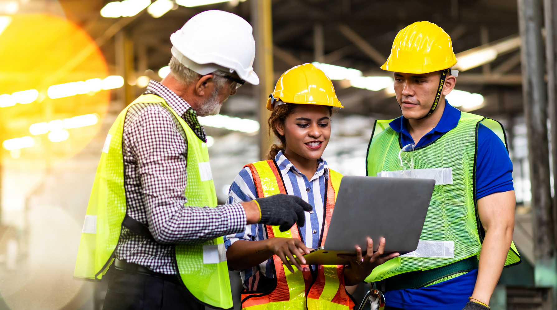 Three workers in hard hats and safety vests collaborating on a laptop at a construction site.